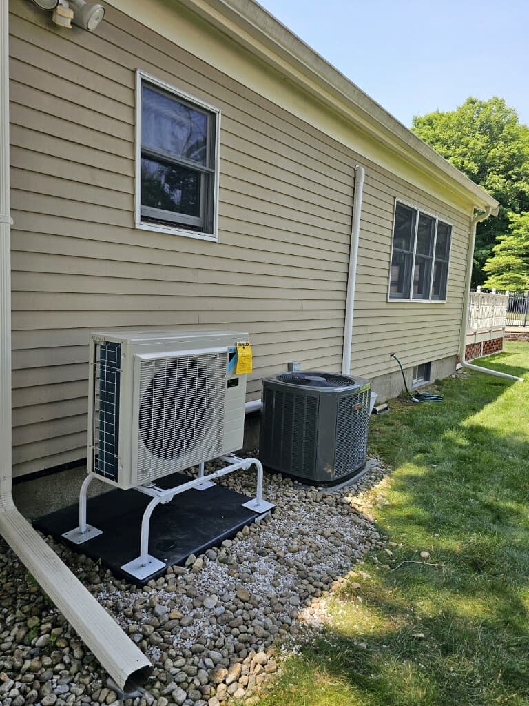 White heat pump and grey air conditioner units outside a house with tan siding.